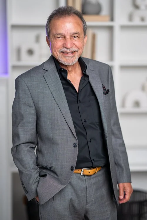 Abid Iftikhar standing in a grey suit and black shirt, smiling